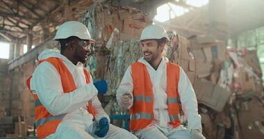 A man with Black skin and a beard in a white protective uniform sits and communicates with his colleague a brunette man with a beard and plays rock-paper-scissors near huge piles of garbage at a video