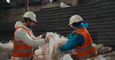 A brunette man with a beard in a white protective uniform and an orange vest together with a brunette girl presses plastic cellophane while working at a waste processing and sorting plant video