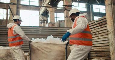 A man with Black skin in a white special uniform and an orange vest lays out and sorts cellophane garbage together with his colleague a man with a beard in a white helmet at a waste processing and video
