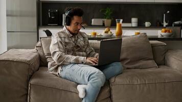 Confident and happy man with Black brunette skin color in black modern headphones plays games on his old laptop while sitting on a modern sofa in a studio apartment video