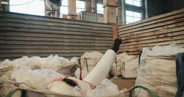 A brunette man in a white protective uniform in a safety helmet and an orange vest jumps into a large pile of recycled plastic cellophane at a waste processing and sorting plant video