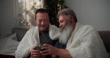 A happy elderly man with gray hair and a lush gray beard is wrapped in a white woolen blanket and drinks hot tea with his brunette boyfriend in a gray gray T-shirt on the sofa in the evening in a video