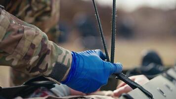 Close up a confident male doctor in a camouflage uniform with blue medical gloves unties the straps on a stretcher for unconscious wounded soldiers during combat operations in the steppe video