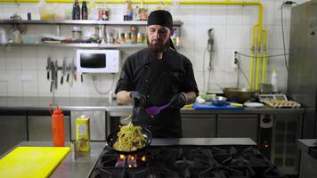 confidente masculino cocinero en un negro uniforme con un negro pañuelo papas fritas vegetales en un gas quemador torneado ellos terminado para incluso Cocinando utilizando un espátula en el cocina en un restaurante video