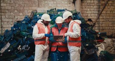 A trio of workers in white uniforms and orange vests sort out their plans on a laptop and communicate while standing near a mountain of plastic garbage at a waste recycling plant video
