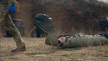 Close-up a man lies unconscious on a green army carrier for the wounded while a medical army soldier drags him along the steppe at an army training ground video