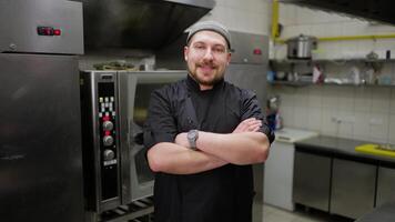 Portrait of a confident professional male chef in a hat and black uniform with a beard who folds his arms on his chest and feels confident in a restaurant kitchen video