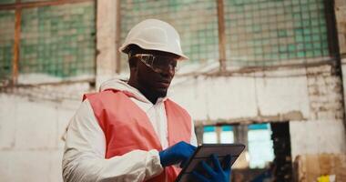 A man with Black skin in a white helmet and an orange vest stands at a huge waste processing plant and writes data into the tablet he is holding in his hands video