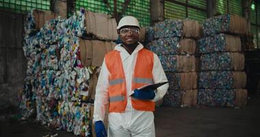 Portrait of a happy man with Black skin in a white uniform and an orange vest who stands near a huge pile of recycled plastic at a waste recycling plant and holds a tablet in his hands video