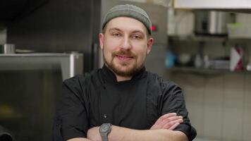 Portrait of a happy chef with a beard in a black uniform who folds his arms on his chest and poses for the camera in the kitchen in a restaurant video