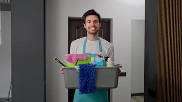 Portrait of a confident brunette janitor guy in a blue uniform and a white T-shirt who holds in his hands a gray and plastic basin filled with a lot of tools for cleaning the apartment in a modern video