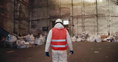 Rear view of a man in a red safety vest and white uniform walking through a tall waste recycling plant. A huge room with polar bears with garbage that will be recycled in a huge plant video
