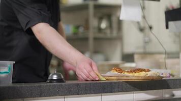 A confident professional chef in a black uniform places a finished dish of cheese flatbread on the table for distribution and rings the bell to signal the readiness of the dish video