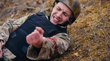 Close-up a soldier in a camouflage uniform and body armor lying wounded in the arm waves his hand to a medic to provide first aid during combat operations in the steppe video