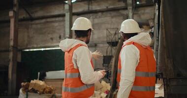 Rear view of a duo of men in special protective clothing and orange vests talking and discussing How to better design plans at their waste recycling plant while recycling waste paper on a conveyor video