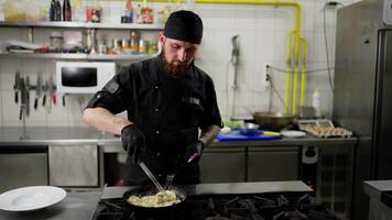 A professional male chef with a beard in a black uniform and with a bandana stirs food in a frying pan while preparing a dish in a restaurant kitchen video