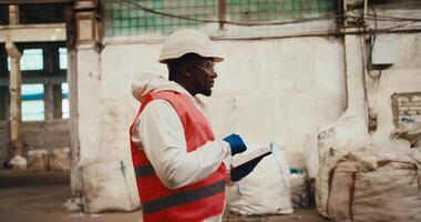 A man with Black skin in a white helmet and a white uniform in an orange vest walks through a waste processing plant with white walls and writes everything down on his tablet. Side view video