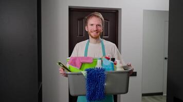 Portrait of a confident blond professional cleaner man in a blue apron who holds a gray plastic basin in his hands which is filled with a large number of cleaning tools while calling a cleaning video