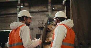 A man with a beard in a white helmet and a white protective uniform stands and communicates with his colleague a man with Black skin in an orange vest near a conveyor belt at a large gray waste video