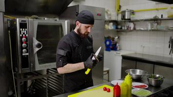Confident male chef in a black uniform with a black bandana and gloves peels an avocado while preparing a salad in the kitchen in a restaurant. Confident chef with beard peels vegetables before video