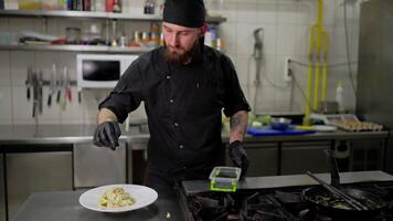 A man with a beard a professional chef in a black protective uniform sprinkles a dish with spices and herbs while preparing it for serving in the kitchen video