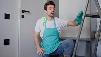 portrait of a confident happy male cleaner in a blue apron throwing a rag over his shoulder and posing while sitting on the floor among cleaning tools and a stepladder in a modern apartment video
