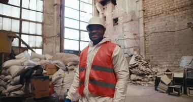 Portrait of a happy confident man with black skin color in a white uniform and orange worker at a garbage and waste processing plant. Male factory worker crosses his arms over his chest video
