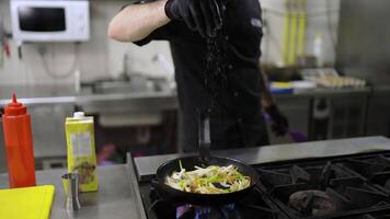 Close-up of a confident man Chef in a black uniform sprinkles vegetables with salt which he fries in a frying pan on the stove in a restaurant kitchen video