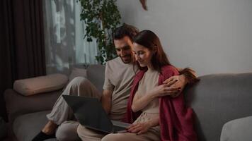 Side view of a happy brunette man with stubble together with his young brunette wife in a red sweater sitting on a gray sofa and watching an interesting movie on a gray laptop in a modern apartment video