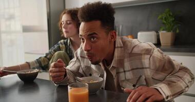 A young man with Black skin color in a cream plaid shirt does not like the cereal with juice that he eats from his bowl and he spits it right there during his breakfast in a modern apartment with his video