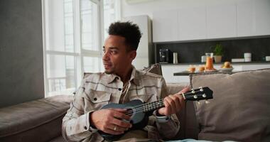 A calm and happy man with Black skin color a young brunette with stubble plays a small musical stringed instrument the ukulele while sitting on a modern sofa in a cream plaid shirt in a modern video