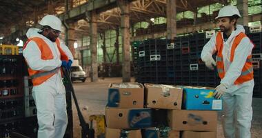 A Black man in a white uniform together with his colleague a man with a beard and an orange vest are trying to put a wheelbarrow with sorted waste in place near boxes with sorted glass waste at a video