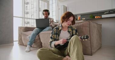 A young girl with a bob hairstyle in a green checkered shirt sits near the sofa on the floor and plays the ukulele while her boyfriend in modern Black headphones works at a gray laptop in a modern video