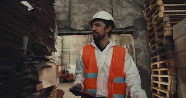 A brunette man with a beard in a white protective uniform in an orange vest walks along the stacks with waste paper and does recounting at a large waste sorting and recycling plant video