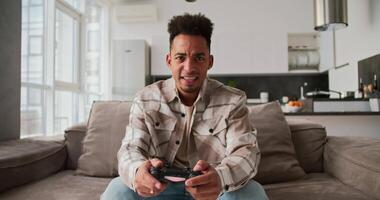 A sad and angry young man with Black skin a brunette with stubble in a cream-colored checkered shirt sits on a sofa in a modern apartment and plays a game using a black joystick and is sad about his video