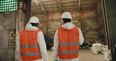 Rear view of two male colleagues in white uniforms and orange vests going out into the premises of a huge waste recycling and sorting plant. A Black man and a brunette man with a beard are talking and video