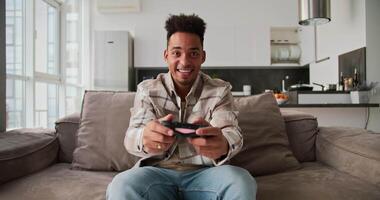 Portrait of a happy young Black-skinned brunette man with stubble who sits on a modern sofa and plays a game and rejoices at his victory using a black joystick in a modern studio apartment video
