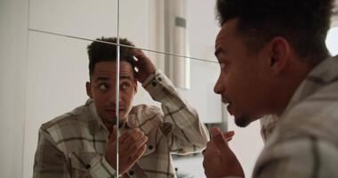 A young Black-skinned brunette man with stubble in a cream-colored checkered shirt preens near a mirror in the hallway in a modern apartment with light walls and good lighting video