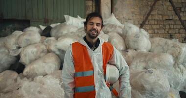 Portrait of a happy and confident brunette man with a beard in a white protective helmet and an orange vest posing against the backdrop of huge piles of recycled and sorted cellophane at a waste video