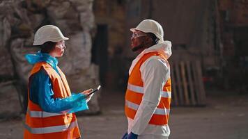 A brunette girl in a blue uniform and an orange vest communicates with a man with Black skin in a white uniform while they are at a large waste processing and sorting plant video