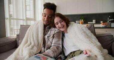Happy young man with Black skin color in a plaid shirt sits with his girlfriend on a modern gray sofa wrapped in a white wool blanket. They read a book and stories in a modern apartment while spending video