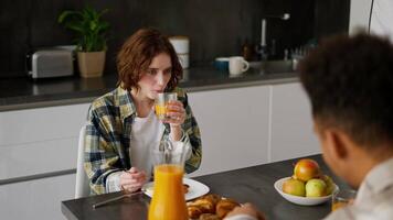 Over the shoulder a happy young adult girl with brown hair and a bob hairstyle in green checkered is having breakfast with her boyfriend with Black brunette skin color during a shared breakfast in the video