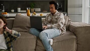 Happy young man with Black skin color in a beige checkered shirt plays computer games on his laptop while his young adult girlfriend with a bob hairstyle does yoga while sitting on the floor in a room video