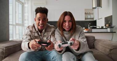 A happy Black-skinned man with a brunette and his girlfriend in a sweater play a game using a joystick. Then the man wins and the girl is sad about her defeat in a modern apartment Sitting on video