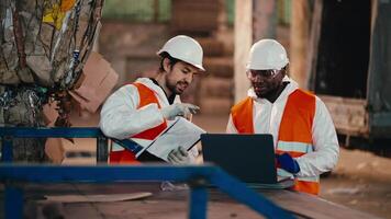 A brunette man with a beard in a white protective uniform in an orange vest talks to his colleague a man with Black skin who works at a laptop while at a large waste processing and sorting plant video