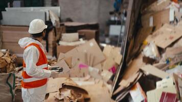 A brunette man with a beard in a white protective uniform and an orange vest stands near a large conveyor with moving waste paper at a large waste paper recycling and sorting plant video