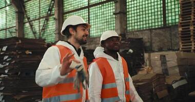 A brunette man with a beard in a white uniform and an orange protective vest walks with his Black-skinned male colleague and communicates at a large waste processing and sorting plant video