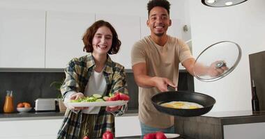 Portrait of a happy young man with Black skin in a cream T-shirt and a young adult girl with a bob hairstyle in a plaid shirt who holds in her hands a salad and a fried egg while preparing breakfast video