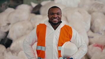 Portrait of a Happy man with Black skin in a white protective uniform who holds in his hands a protective white helmet and an orange vest on it. He stands near large bags of cellophane and video