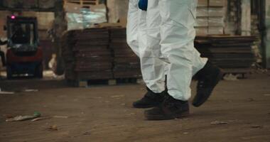 Close-up shot of two engineer employees in white protective uniforms and black shoes walking along a plant for recycling waste paper and plastic video